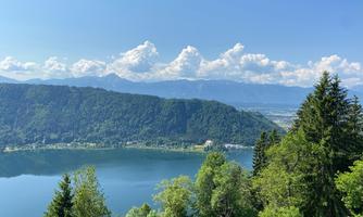 A beautiful view of a clear lake, surrounded by trees and mountains. The sky is blue with some clouds.