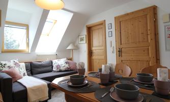 A cozy living space with a sofa, a dining table, and wooden details. The large windows provide plenty of daylight.
