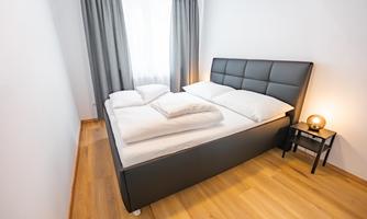 A modern bedroom with a comfortable double bed and bright curtains. The floor is wooden, and a nightstand with a lamp complements the decor.