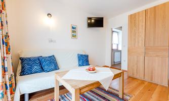 A cozy living room with a bright sofa and colorful cushions. A wooden table with a fruit basket and a TV on the wall create an inviting atmosphere.