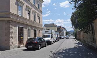 A quiet street with parked cars and a large building on the left side. The sky is blue with white clouds and there are trees along the roadside.