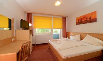 A cozy room with a double bed and a desk. Large windows with yellow blinds and a TV give the space an inviting atmosphere.