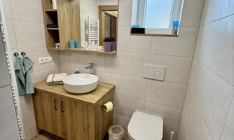 A modern bathroom with a wooden washstand and a round sink. The walls are brightly tiled and there is a window that lets in daylight.