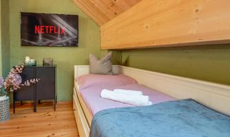 A cozy bedroom with a single bed and a television showing Netflix. The walls are green, and warm wooden elements create an inviting atmosphere.