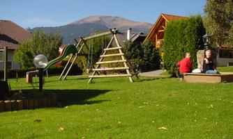 A beautiful garden with a slide and a climbing frame. In the background, there are mountains and two people are sitting relaxed in the grass.