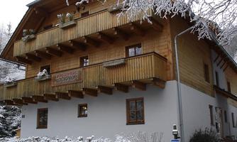 A cozy wooden house in winter with white snow drifts. The balcony has wood-decorated railings and plants.