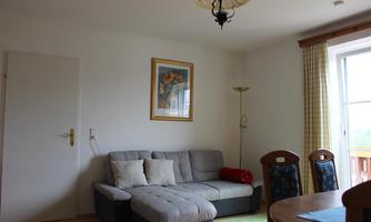 A cozy living room with a gray sofa, a table, and chairs. There is a picture hanging on the wall and large windows with yellow curtains.