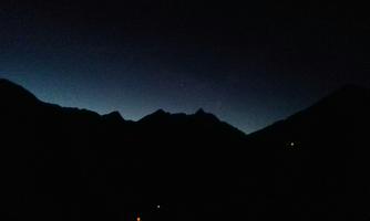 A gloomy mountain landscape at night. In the horizon, a faint light illuminates the silhouettes of the mountains.
