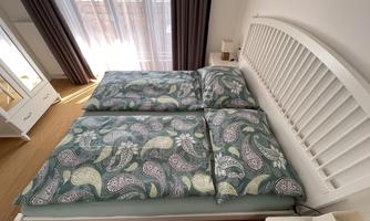 A bright bedroom with a white bed and colorful duvet covers. Soft rays of light fall on the wooden floor in front of the window.