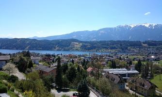 A picturesque view of a lake surrounded by hills and mountains. In the foreground, green meadows and residential houses are visible.