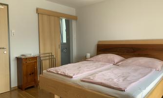 A cozy bedroom with a double bed and two pillows. The walls are painted light, and a wooden wardrobe is visible in the room.