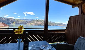 A cozy balcony with a view of a lake and the mountains. On the table are a wine glass, a plate, and a flower decoration.