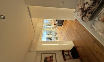 A modern bedroom with wooden floors and large windows. In the background, there is a seating area with a view outside.