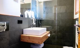 A modern bathroom with a black tile wall and a floating sink. There is a shower and towels nearby.