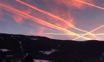 A spectacular sunset over the mountains, with clear contrails coloring the sky in warm shades of red. The landscape appears calm and picturesque.