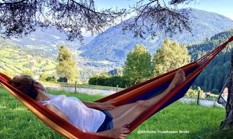 A person lies relaxed in a hammock in nature. In the background, gentle hills and trees can be seen.