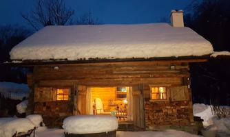 A cozy cabin in the snow, illuminated by warm light. The eaves are thickly covered with snow.