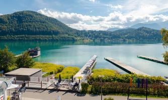 A tranquil lake with clear water and green shores. In the background, gentle hills and a blue sky with a few clouds can be seen.