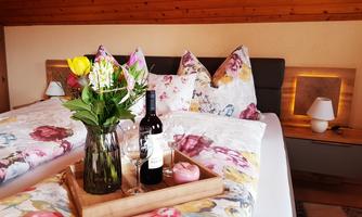 A cozy bedroom with blooming pillows and a romantic atmosphere. On the bed, there is a tray with wine, glasses, a flower, and a candle.