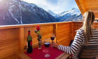 A cozy balcony with a view of snow-covered mountains. One person is sitting there with a glass of red wine, a bouquet of roses, and a bottle of wine on the table.