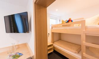 A cozy children's room with a bunk bed and a TV on the wall. There is a window with natural light and toys on the upper bed.