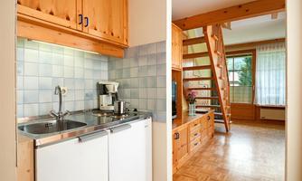 A modern kitchen with wooden furniture and a small dining area is visible. In the background, there is a bright living space with stairs and large windows.