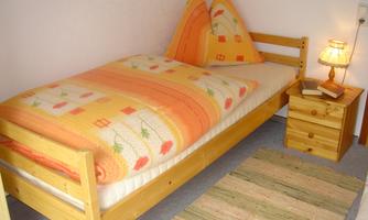 A simple bedroom with a single bed adorned with an orange duvet and pillows. Next to the bed is a nightstand with a lamp and a book.