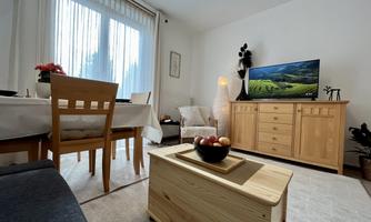 A cozy living room with a wooden table and chairs. In the background, there is a television displaying a landscape view, and large windows provide daylight.
