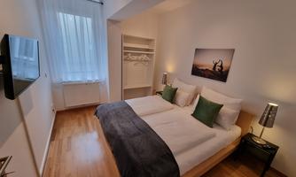A modern bedroom with a large double bed and green pillows. There is a bright window front, a television, and simple furniture.