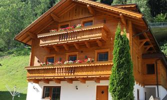 A traditional wooden house with several balconies and colorful flowers. It is located in a green environment with trees in the background.