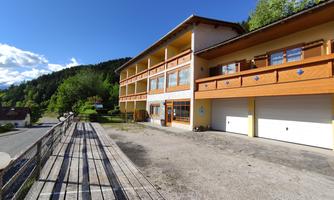 A modern building with wooden cladding and large windows stands in a quiet environment. In the background, trees and a blue sky can be seen.
