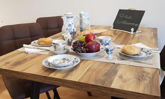 A cozy table with breakfast, consisting of rolls, fruit, and dishes. A small sign welcomes the guests.
