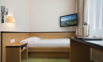 A simple hotel room with a single bed, desk, and a wall-mounted television. The room has large windows and a bright, friendly atmosphere.