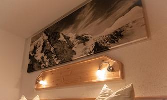 A cozy room with a wooden bed and cushions. Above the bed hangs a large black-and-white picture of snow-covered mountains.