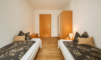 A modern bedroom with two single beds and stylish pillows. The room is bright and cheerful with wooden flooring and simple furnishings.