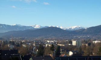 An impressive landscape with snow-covered mountains in the background. In the foreground, the roofs of a city with trees can be seen.