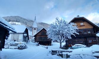 A snowy village with traditional wooden houses and a church tower. The surroundings are surrounded by mountains and a clear sky.