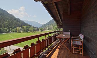 A balcony with a wooden railing and a table overlooks a green meadow and the surrounding mountains. The sky is clear and sunny.