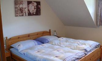 A cozy bedroom with a double bed and blue blankets. Pictures hang on the wall and a window provides daylight.