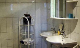 A simple bathroom with white tiles and two sinks. There is a mirror and a shelf with towels.