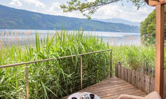 A beautiful view of a lake, surrounded by green reeds. On the terrace, there is a small table with tea cups and a cozy armchair.