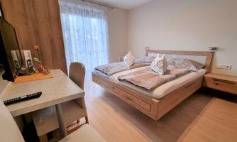 A bright, modern bedroom with a double bed and large windows. The room is simply furnished and includes a desk and a television.