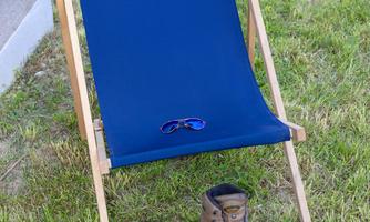 A blue deck chair with the inscription "Hochrindl" is on a green meadow. Next to the chair are hiking boots.