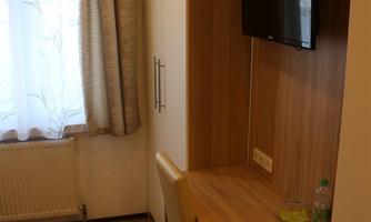 A small, modernly furnished room with a desk, a television, and a window with curtains. The walls are wood-colored, and the floor is laid with laminate.