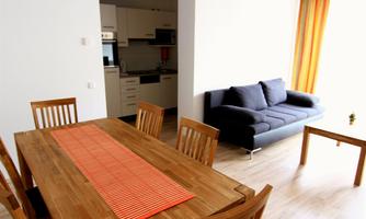 A bright living room with a wooden dining table and a gray sofa. The kitchen in the background is modern and well-equipped.