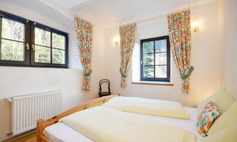 A bright bedroom with two single beds and decorative curtains. The room has large windows that let in plenty of daylight.