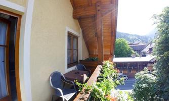 A sunny balcony with a cozy seating area and plants. In the background, green hills and additional buildings can be seen.