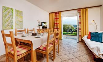 A cozy living area with a wooden table, chairs, and colorful curtains. Through the door, you can see a beautiful view of nature.