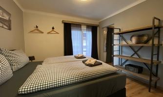 A modern bedroom with two beds and checkered bed covers. The walls are neutrally designed and an open shelf stands in the room.