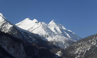 An impressive mountain landscape with snow-capped peaks and a clear blue sky. The surrounding valleys are covered with coniferous forests.